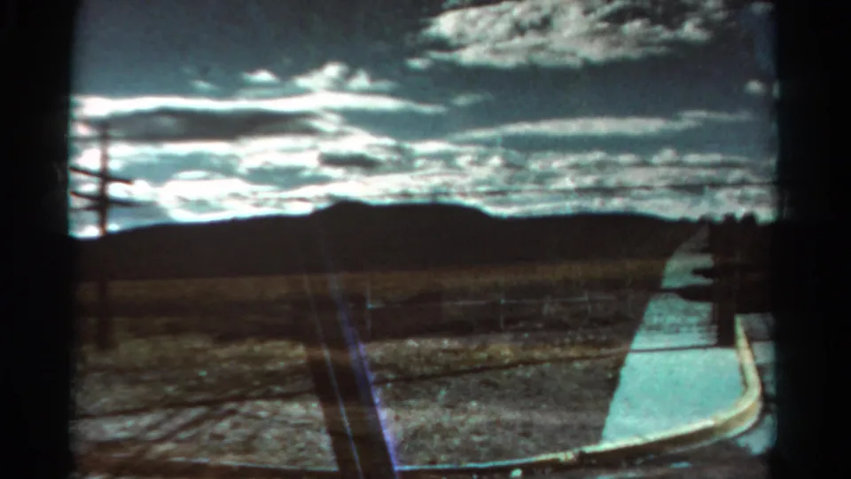 1967 Colorado 8mm frame showing dry open terrain, distant low mountains, fence line, utility pole, and bright clouds.