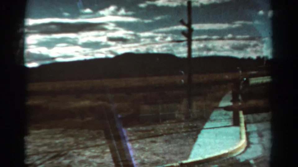1967 Colorado home-movie view of a pale roadside edge beside dry terrain, fence posts, utility pole, and low mountains under clouds.