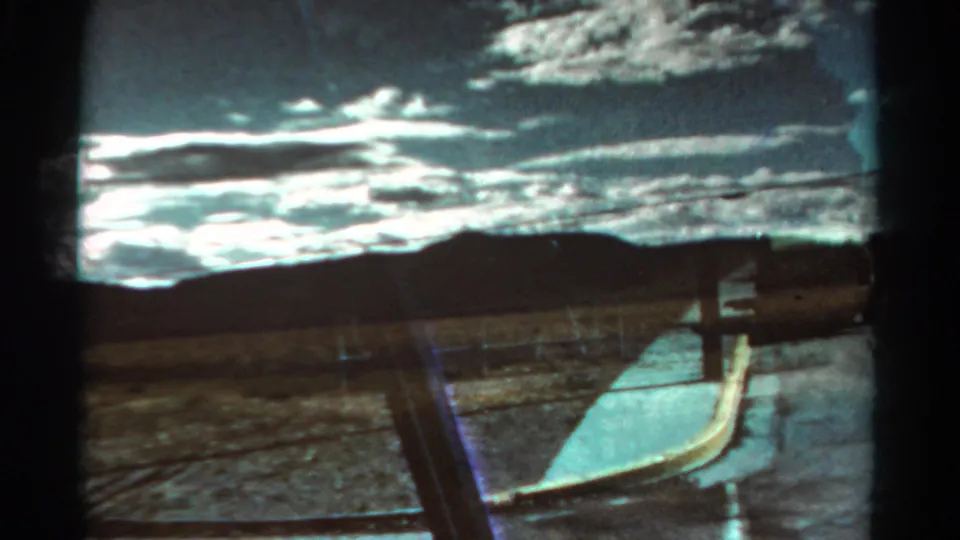 1967 Colorado 8mm frame with dry roadside ground, a fence line, pale path, distant hills, and high-contrast cloud cover.