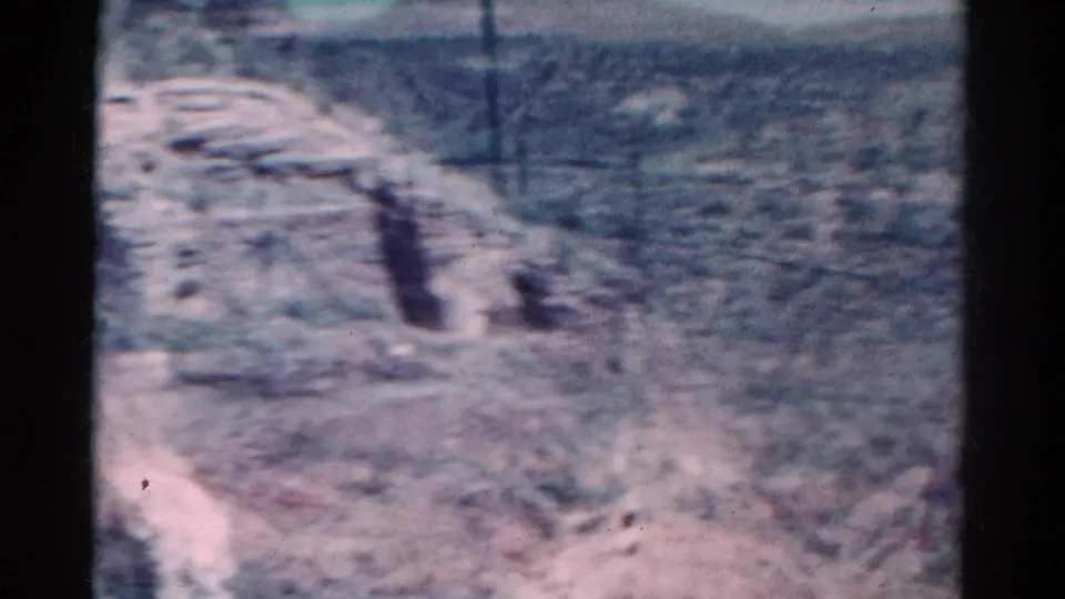 Faded 1967 Colorado 8mm frame showing arid ground, sparse brush, uneven terrain, and a low distant slope.