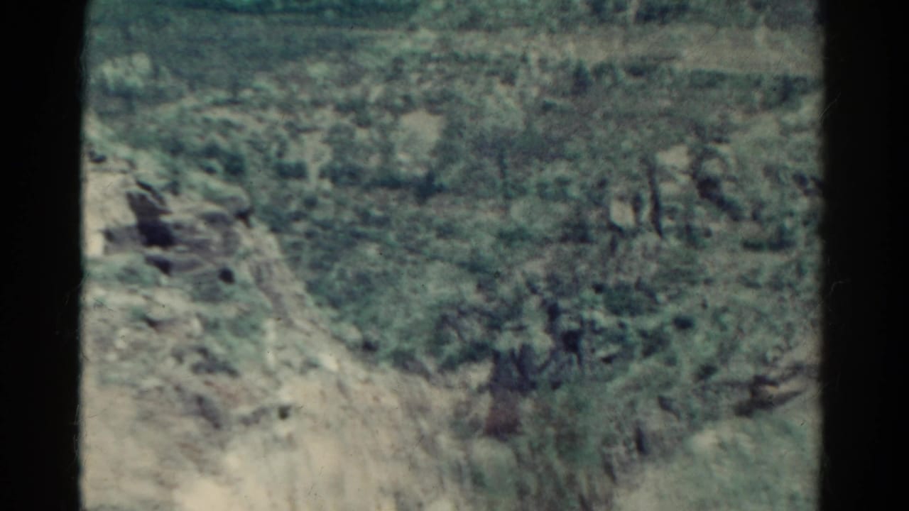 1967 Colorado home-movie frame of a pale rocky slope, scrub growth, and small trees on uneven canyon terrain.