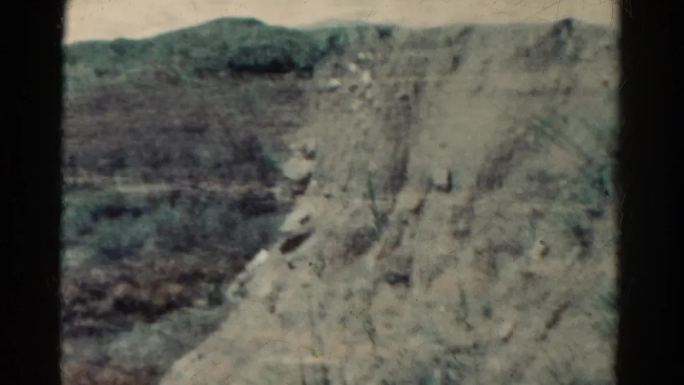 1967 8mm Colorado travel frame showing a pale rocky cliff, scrub vegetation, and distant hills in soft focus.