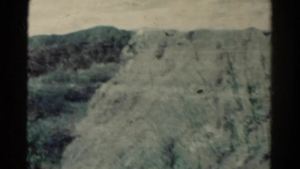 Soft 8mm frame of Colorado mountain terrain, showing a blurred rocky slope, brush, and a low ridge in the distance.