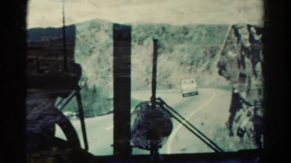 1967 Colorado road-travel frame with a white car on a curving canyon road, viewed through vehicle interior bars and a small fan.