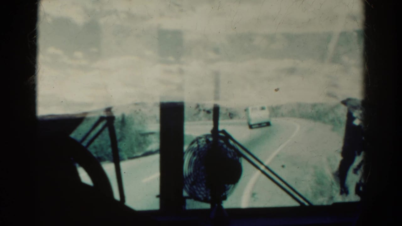 View through a dirty windshield toward a curving road with a white van ahead and a small fan on the dash.