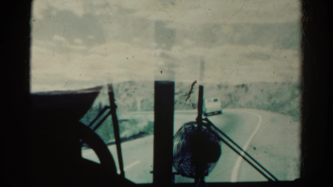 Vehicle interior view through the windshield with a dashboard fan, wiper arms, road curve, and distant van.