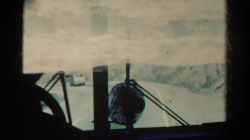 Inside-vehicle view from 1967 Colorado footage, with a dashboard fan silhouetted against a hazy windshield and van on the road ahead.