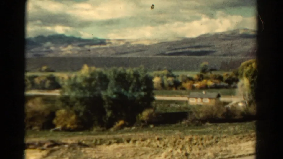 A 1967 Colorado rural view shows fields, trees, small houses, distant mountains, and a cloudy sky on 8mm film.