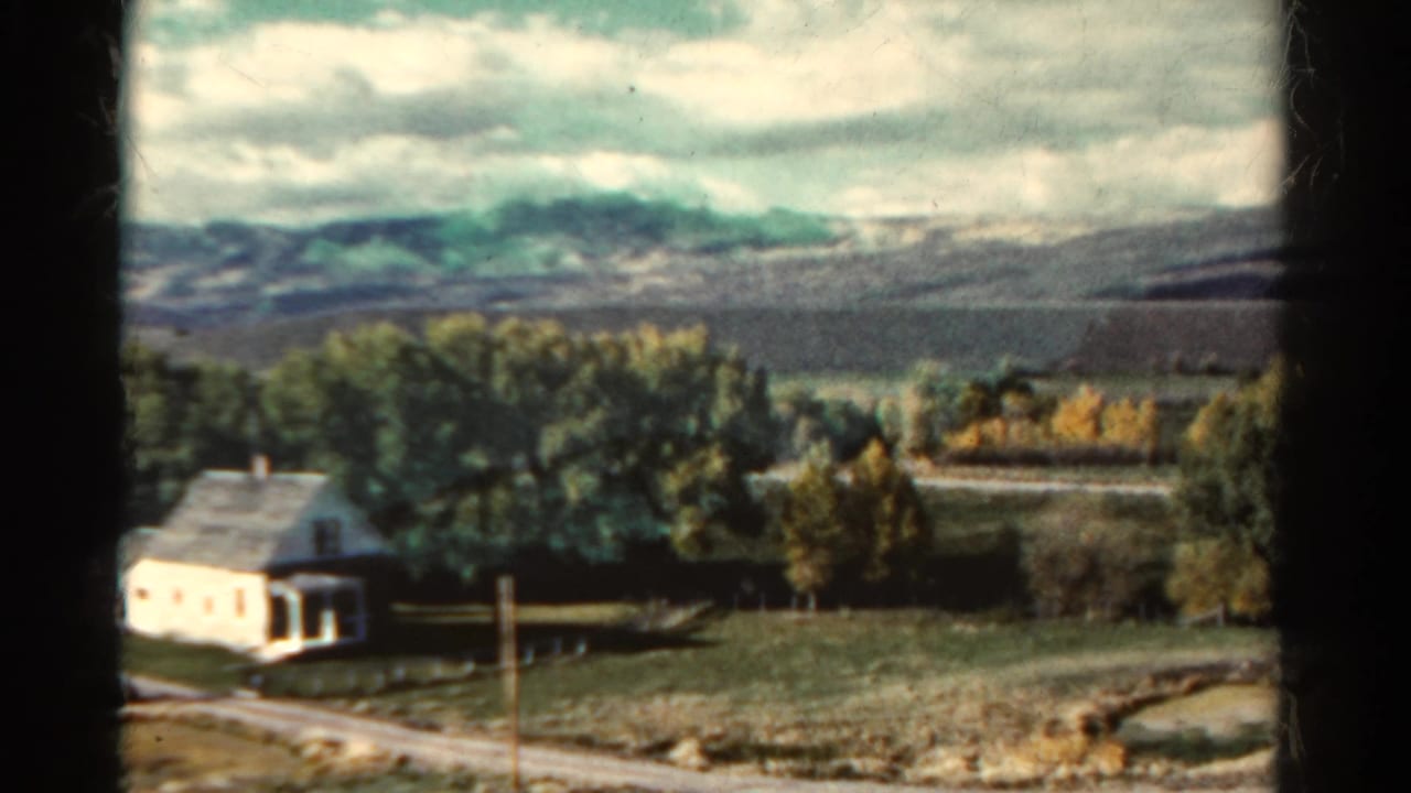 White farmhouse beside trees with fields and distant mountains under cloudy sky.