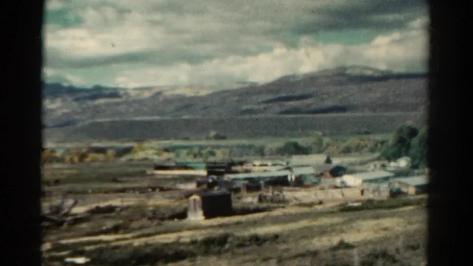 1967 Colorado home-movie frame with a small dark roadside structure, scattered rural buildings, fields, and hills beyond.