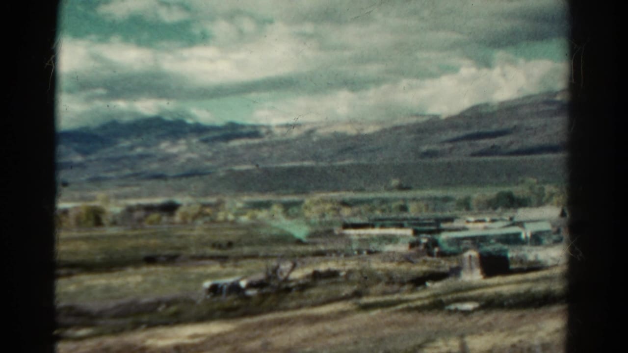 Train-window view of rural buildings, open fields, cloudy sky, and distant mountains.