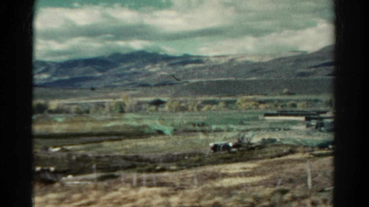 Rural valley with scattered structures, open ground, mountains, and cloudy sky.