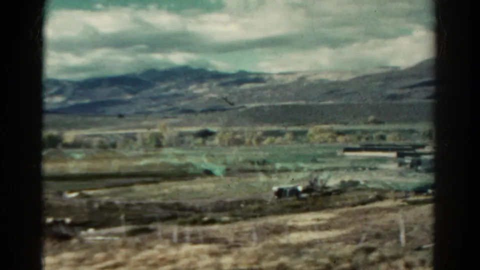 1967 Colorado train-view frame with scattered rural structures, broad open land, mountain ridges, and overcast sky.