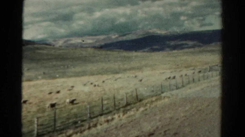 1967 Colorado train-view frame with a fence line, grazing animals, dry field, distant mountains, and heavy cloud cover.
