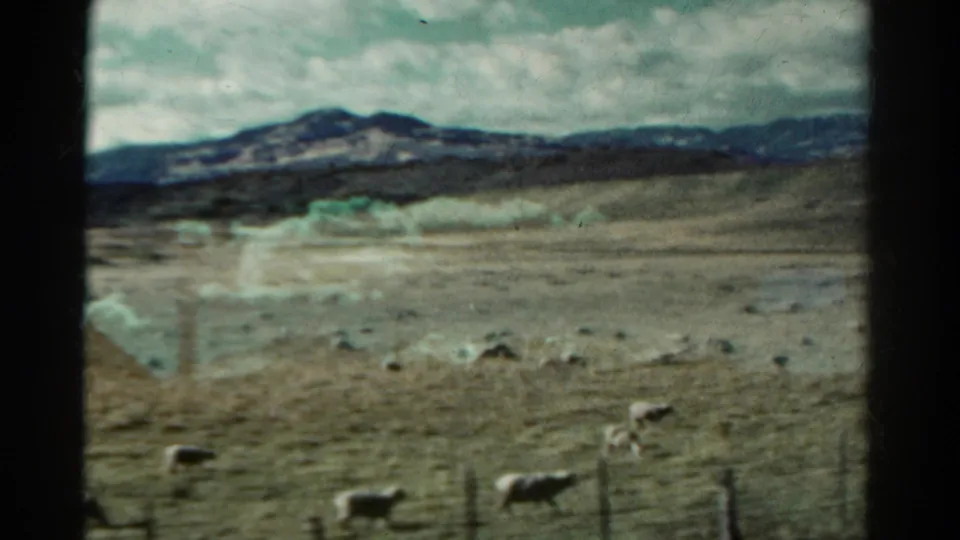 1967 Colorado 8mm still with sheep in the foreground, dry rolling pasture, and a dark mountain ridge under heavy clouds.