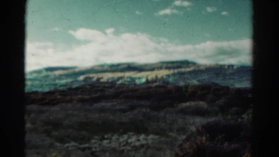 1967 Colorado landscape frame with rolling hills, cloud cover, dark scrub foreground, and a small aircraft-like speck overhead.