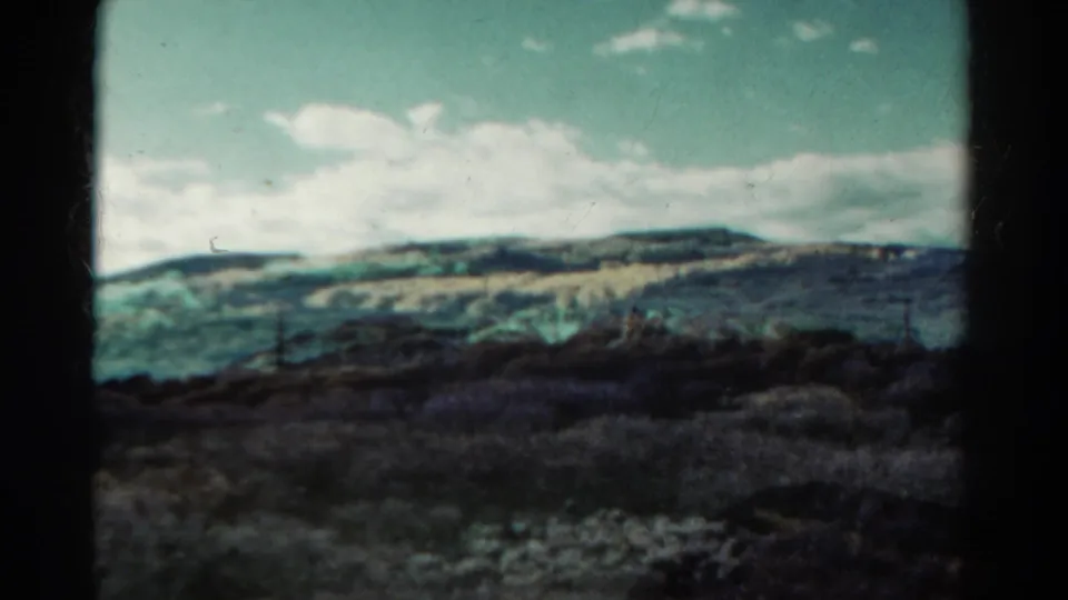 1967 Colorado home-movie frame holding on distant hills beneath bright clouds, with scrub vegetation in front.