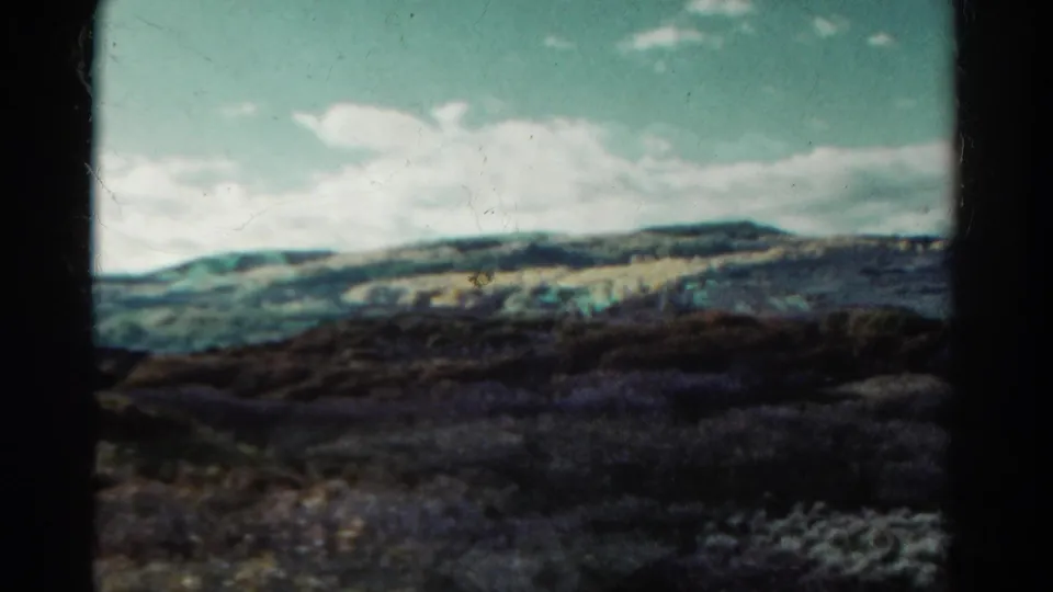 1967 Colorado 8mm frame showing rolling foothills, dark shrub-covered foreground, and scattered white clouds in a pale blue sky.