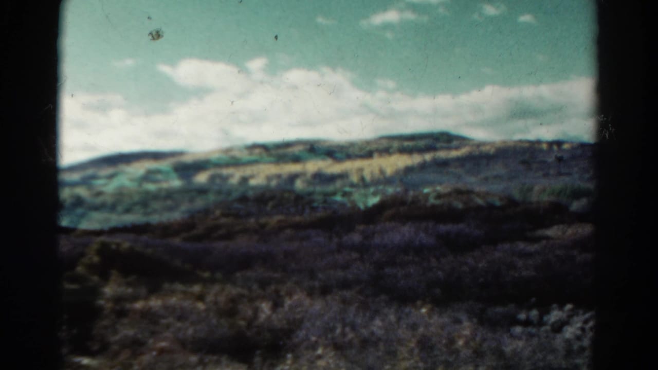 1967 Colorado home-movie frame with distant foothills, low shrubs, and clouded sky, seen through dark 8mm frame edges.