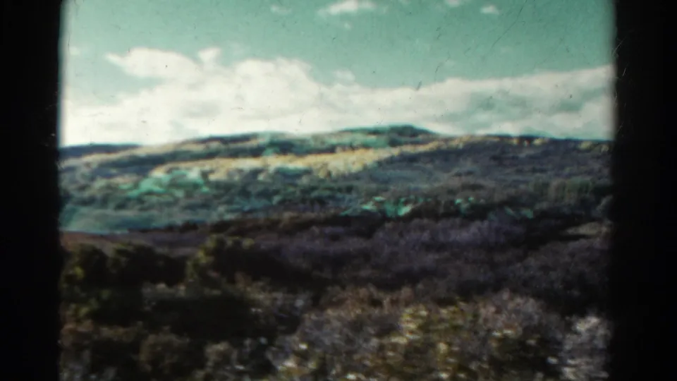 Darker 1967 Colorado 8mm frame showing shrubs and a distant foothill ridge under clouds with a blue-green cast.