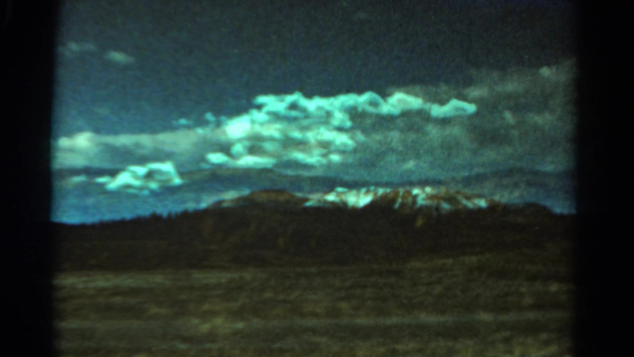 Open plain seen from a train window with dark hills, distant snowy mountains, and bright clouds.