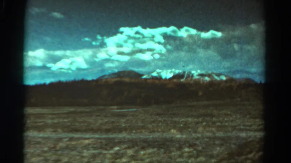 A 1967 8mm train-window frame shows open Colorado terrain, a dark hill band, distant snow-capped mountains, and heavy turquoise cloud color.