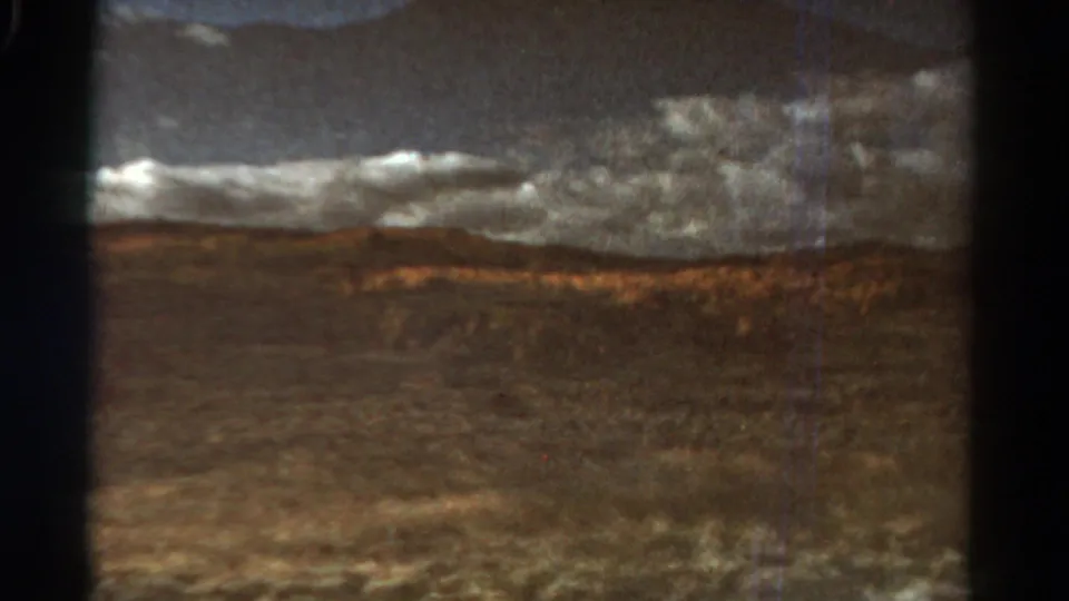 The final 1967 Colorado train-window frame shows a warmer, grainy view of open ground, low hills, and thick clouds with no people or buildings visible.