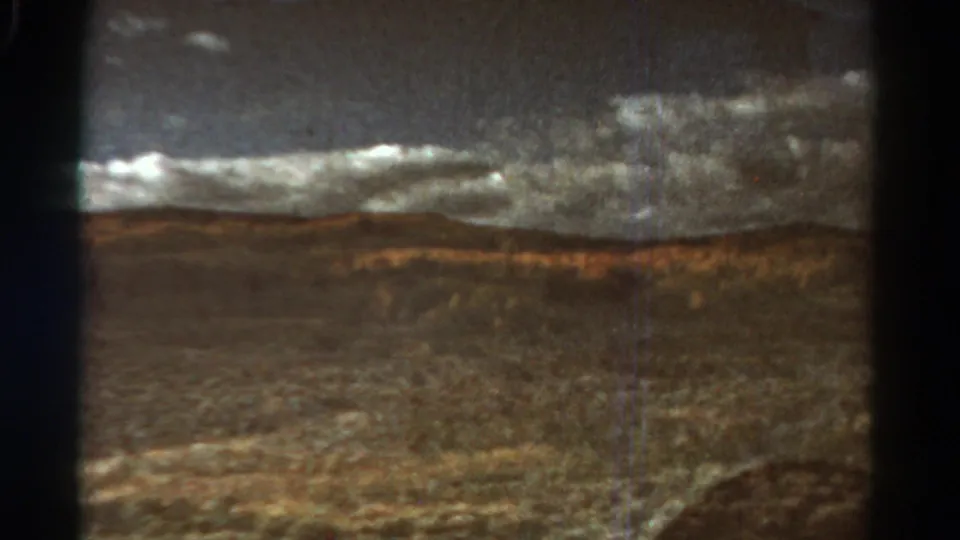 1967 Colorado 8mm frame showing a train-window view across brown valley terrain toward low hills beneath heavy grey clouds.