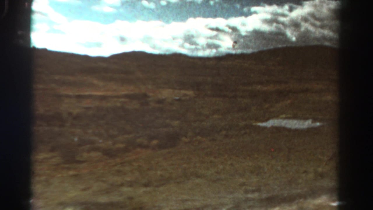 Dry open ground with a small puddle, distant low hills, and bright clouds.
