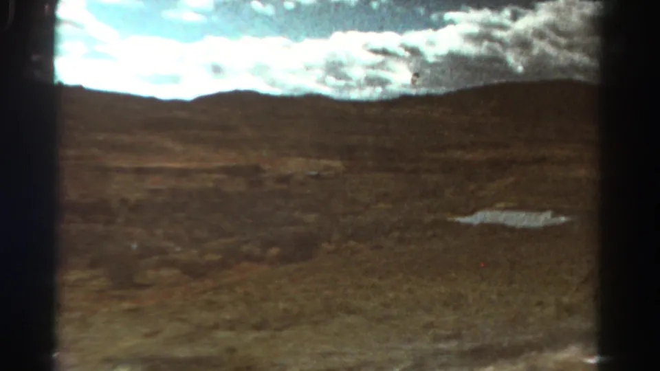 1967 Colorado 8mm frame showing dry open terrain, a shallow puddle, low hills, and a bright clouded sky.