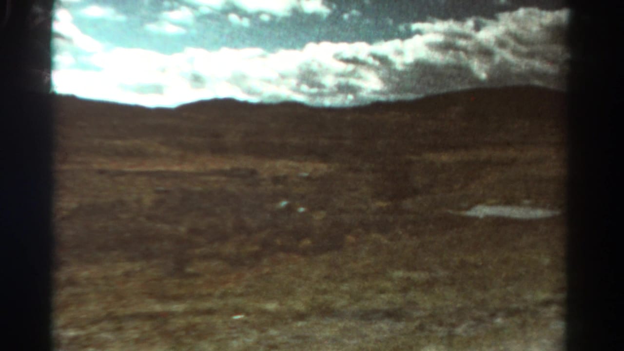 Open brown terrain with a puddle at right, low hills, and clouds across the sky.