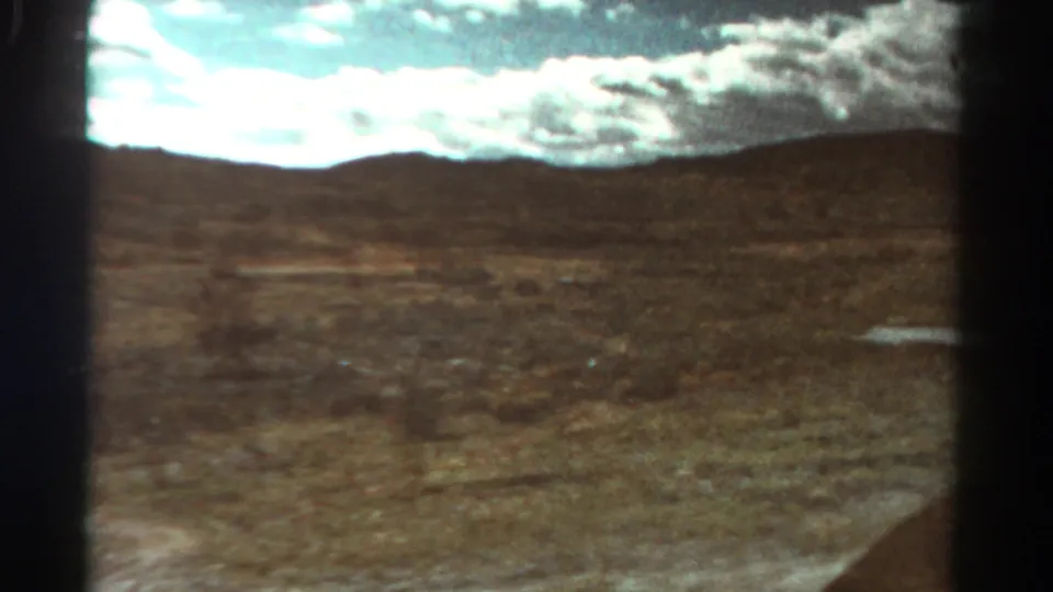 1967 Colorado 8mm frame with soft, blurred dry terrain, distant hills, bright clouds, and a small puddle at right.