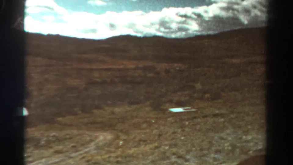 1967 Colorado 8mm frame showing dry gravelly ground, distant hills, bright clouds, and a small light object in the foreground.