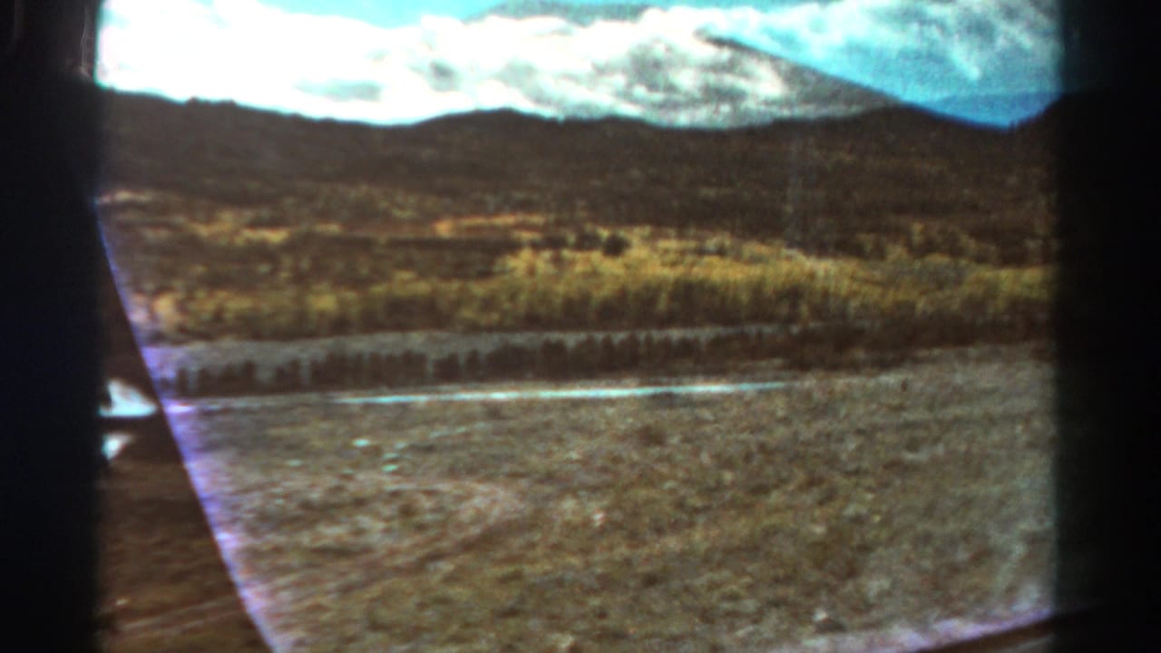 1967 Colorado 8mm view from inside a car, looking past dry terrain, a narrow watercourse, low hills, and distant mountains.