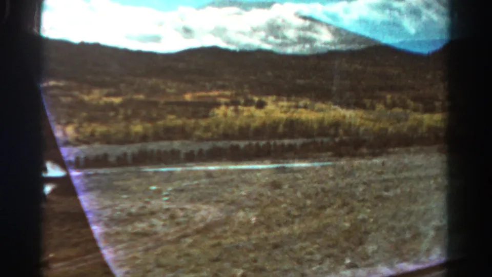 1967 Colorado 8mm view from inside a car, looking past dry terrain, a narrow watercourse, low hills, and distant mountains.