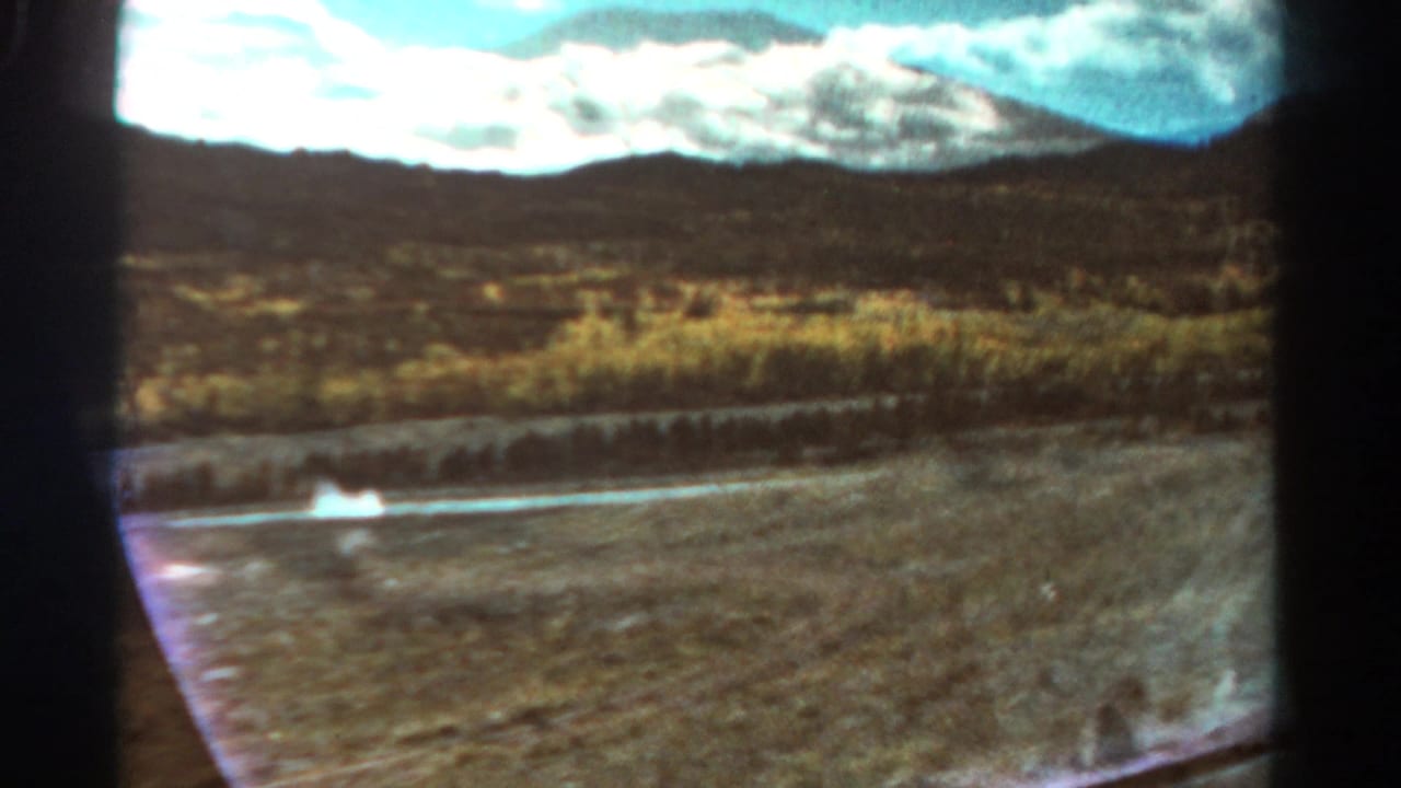 Dry roadside landscape with water, scrub vegetation, hills, and mountains seen through a car window.