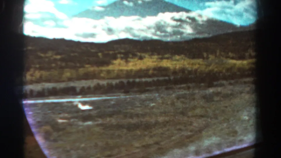 1967 Colorado home-movie frame from a car, showing dry open ground, roadside water, scrub vegetation, hills, and clouded mountains.