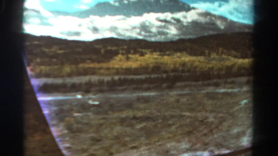 Colorado 1967 8mm frame from inside a moving car, with arid roadside terrain, scrub growth, water, hills, and distant mountains.
