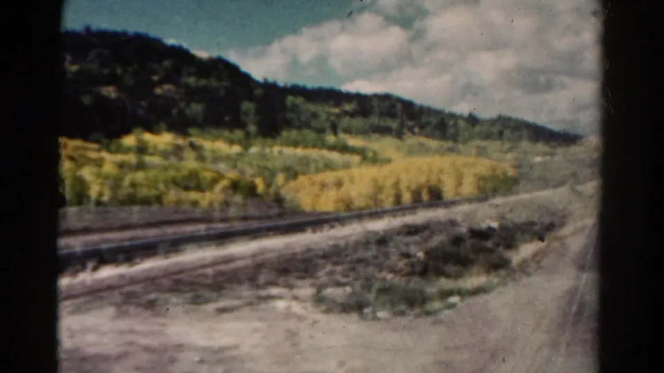 1967 Colorado roadside frame showing autumn trees, a rail line, rough ground, and dark evergreen hills under a cloudy sky.