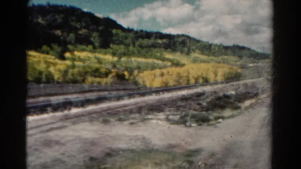 1967 Colorado 8mm frame showing railroad tracks beside golden aspen groves, rough ground, evergreen hills, and cloudy sky.
