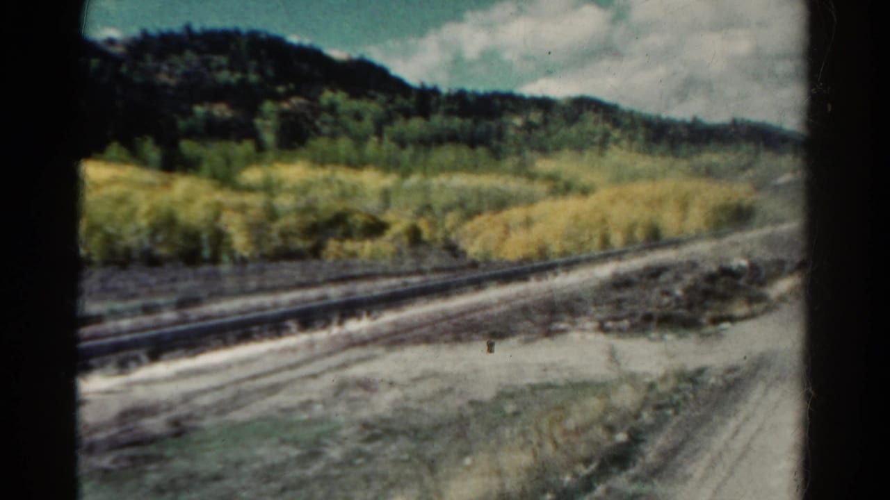 1967 Colorado train-window view with rails, pale dirt, brush, golden aspen stands, and dark wooded hills in soft 8mm focus.