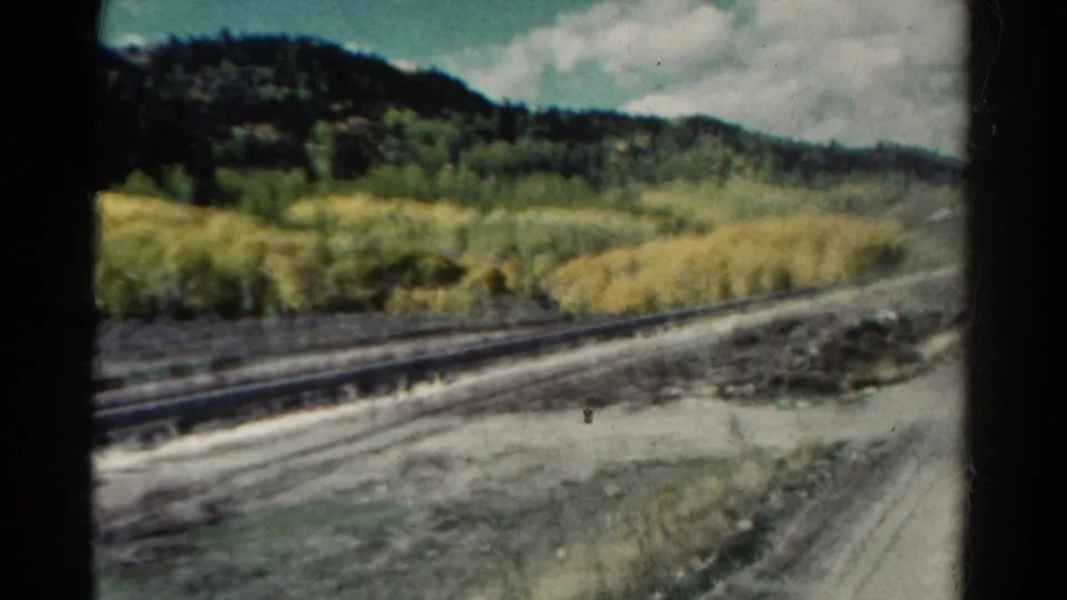 1967 Colorado train-window view with rails, pale dirt, brush, golden aspen stands, and dark wooded hills in soft 8mm focus.