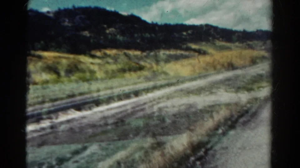 Soft 8mm frame from a moving train in Colorado, with tracks, grasses, golden tree cover, dark hillside, and motion blur.