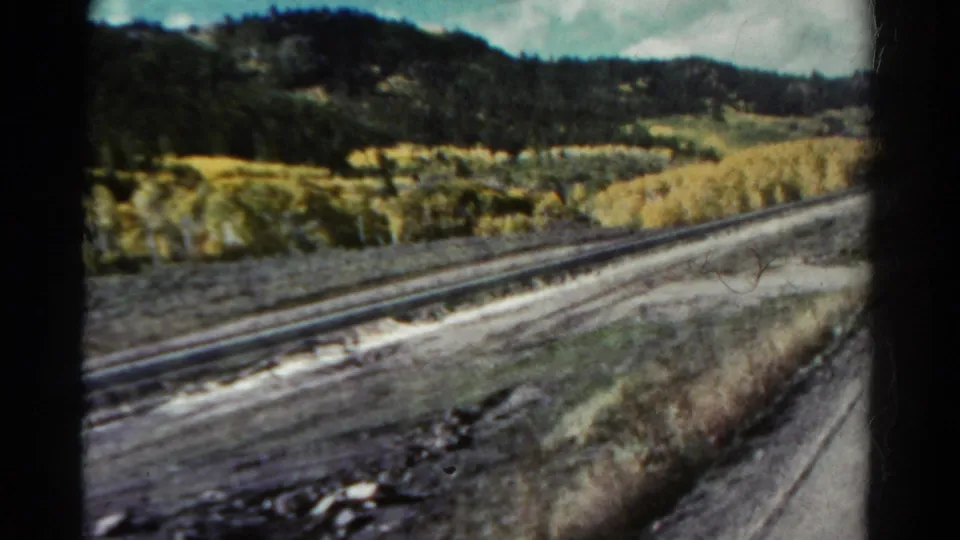 1967 Colorado 8mm frame showing rails beside rocky trackside ground, dry grasses, golden aspens, evergreen slopes, and blue sky.