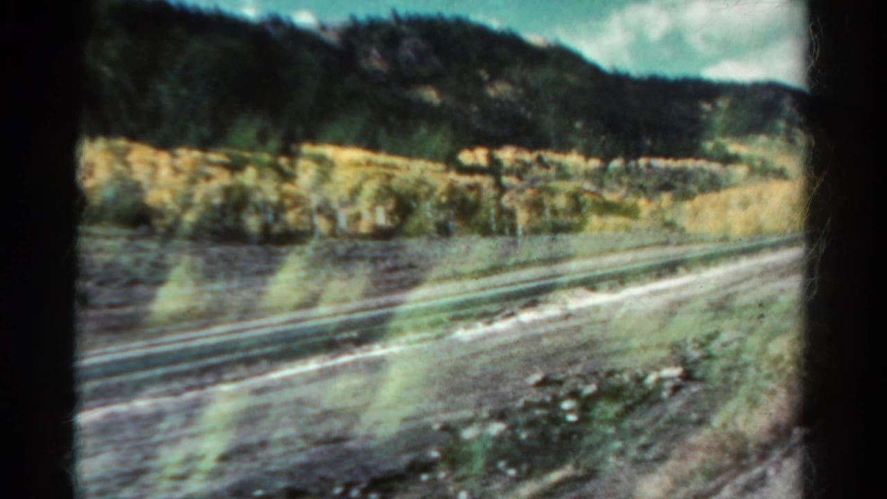 Moving 8mm view of a Colorado rail corridor, with tracks, roadside ground, yellow grasses, and dark hills.