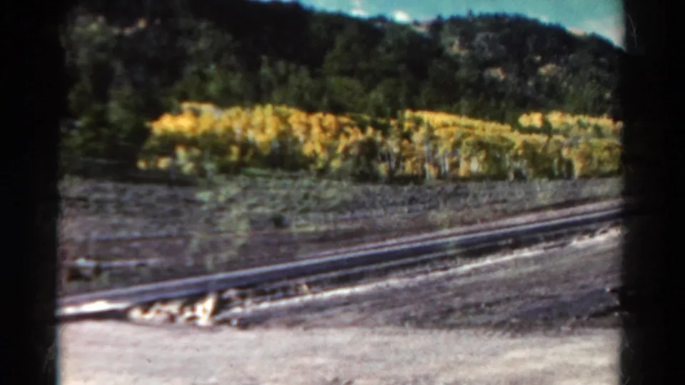 1967 Colorado travel frame with rail tracks in the foreground, dry vegetation, and a dark wooded hillside beyond.
