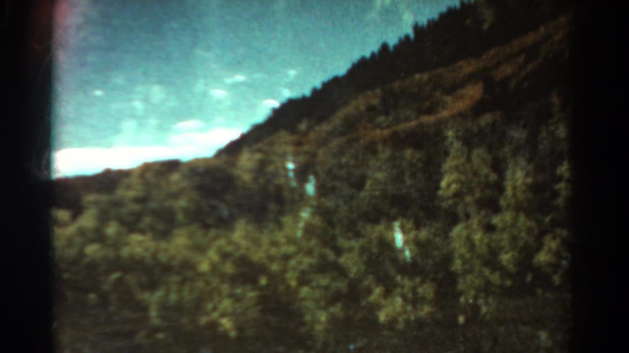 1967 Colorado mountain landscape with evergreen-covered slopes, a bright clouded sky, and visible 8mm color softness.