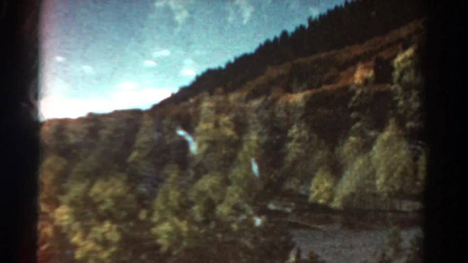 1967 Colorado 8mm frame with a sloped, tree-dotted mountainside, dark frame edge, and partly clouded open sky.