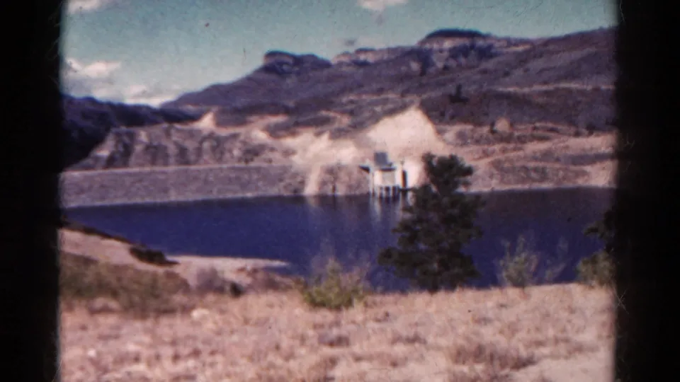 1967 Colorado 8mm view across a reservoir toward rocky mountain slopes, a dam face, and a small waterside structure.