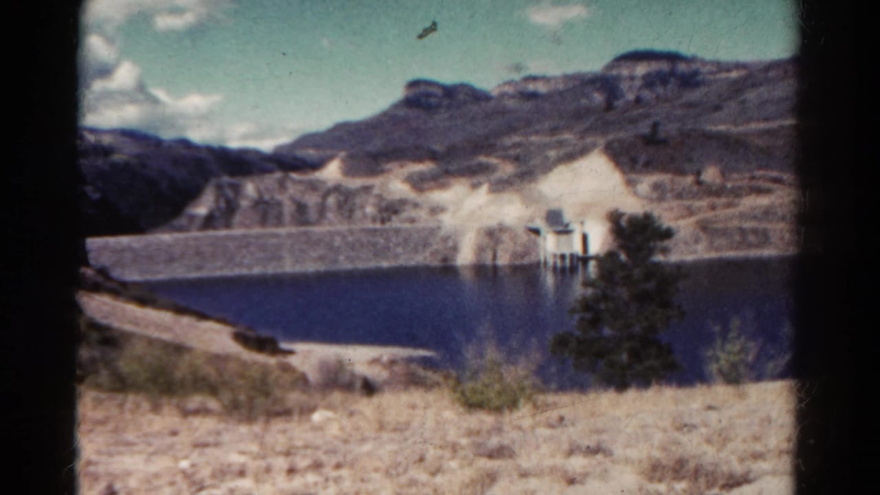 Mountain lake below rocky slopes with a small structure and evergreen trees in the foreground.